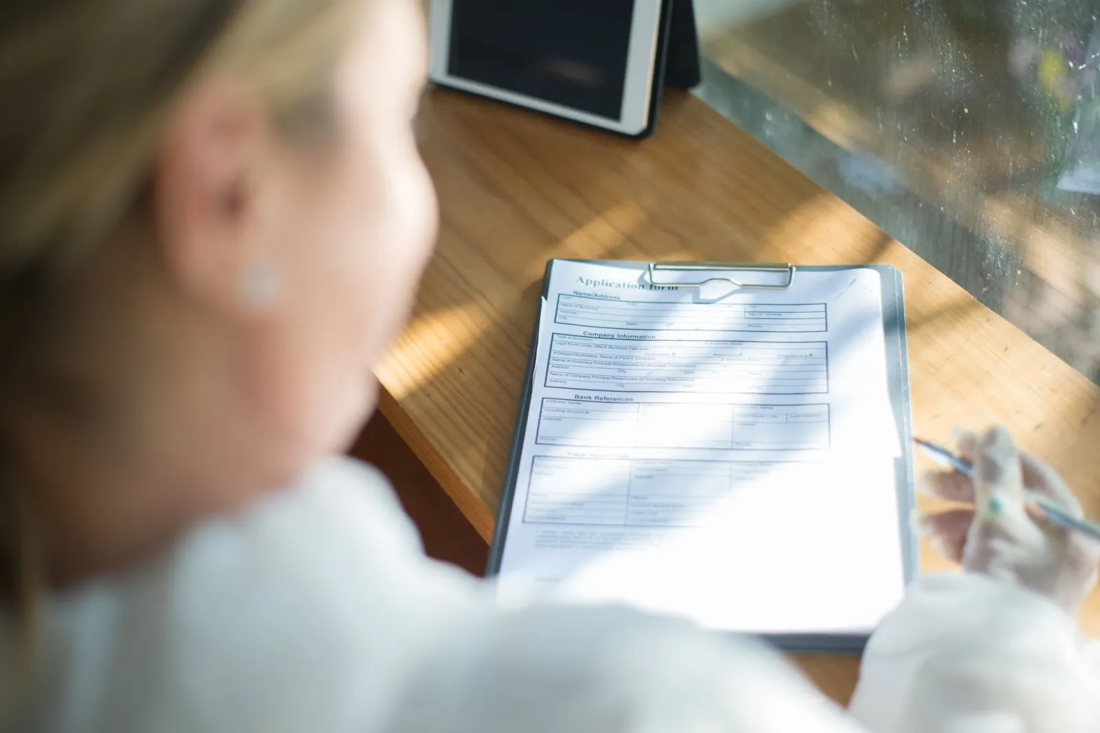 A woman fills out an application form with a pen at a sunlit wooden desk.